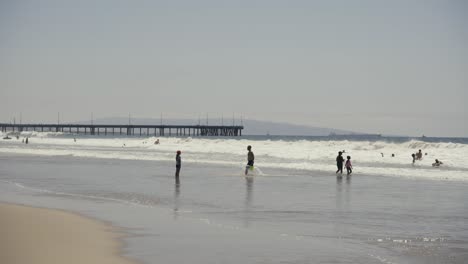 people and families swimming and walking on venice beach with a pier in the background in the afternoon