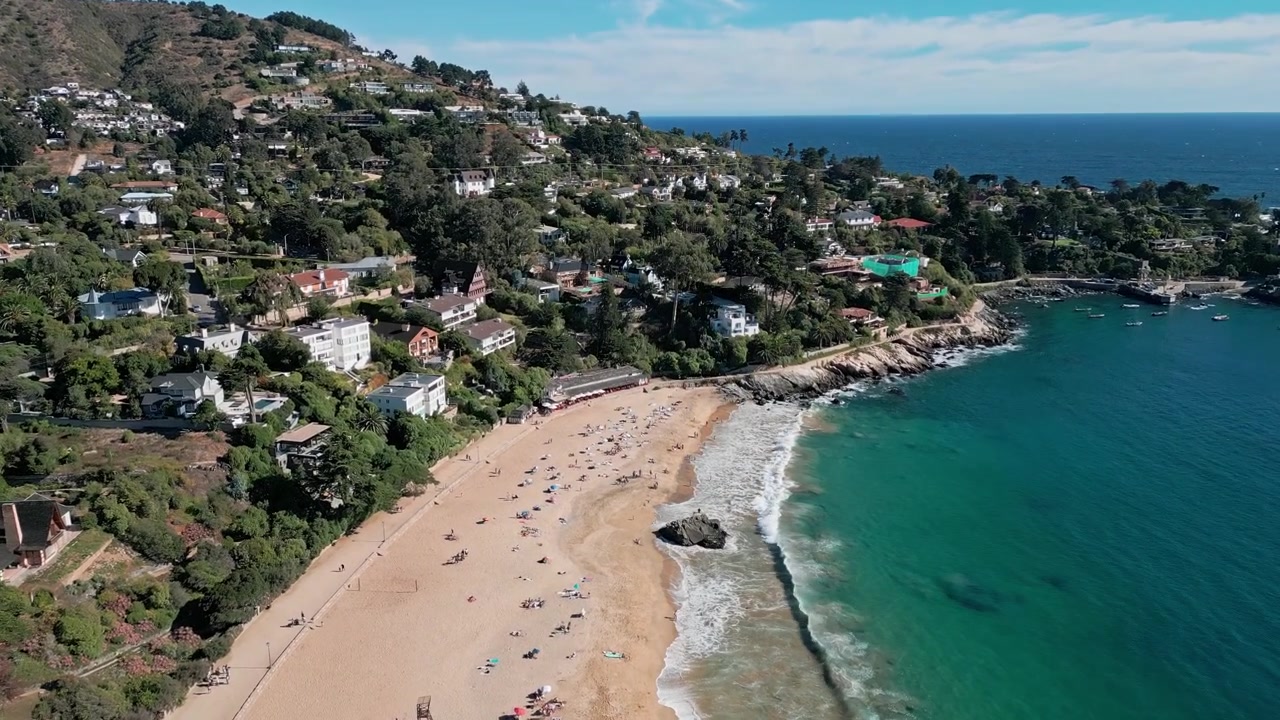 Lenta ripresa in picchiata su un'esclusiva collina residenziale che rivela una spiaggia sabbiosa estesa con bagnanti, calette rocciose e l'orizzonte aperto del Pacifico. Zapallar, Cile.