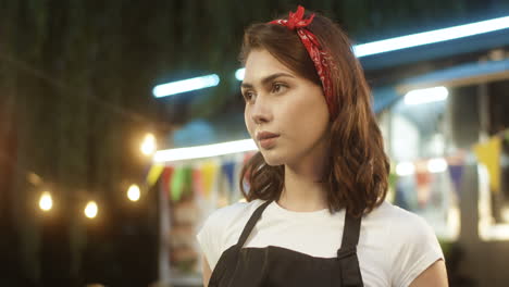 Portrait-Of-Attractive-Joyful-Young-Girl-In-Apron-Smiling-Happily-To-Camera-Outdoor-With-Small-Food-Truck-On-Background