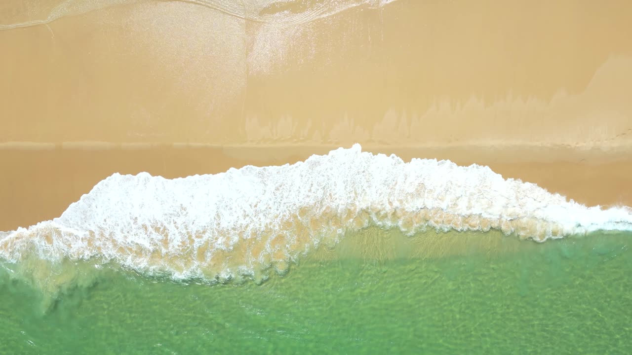 Bird's eye drone view over waves breaking on tropical sand beach in slow motion