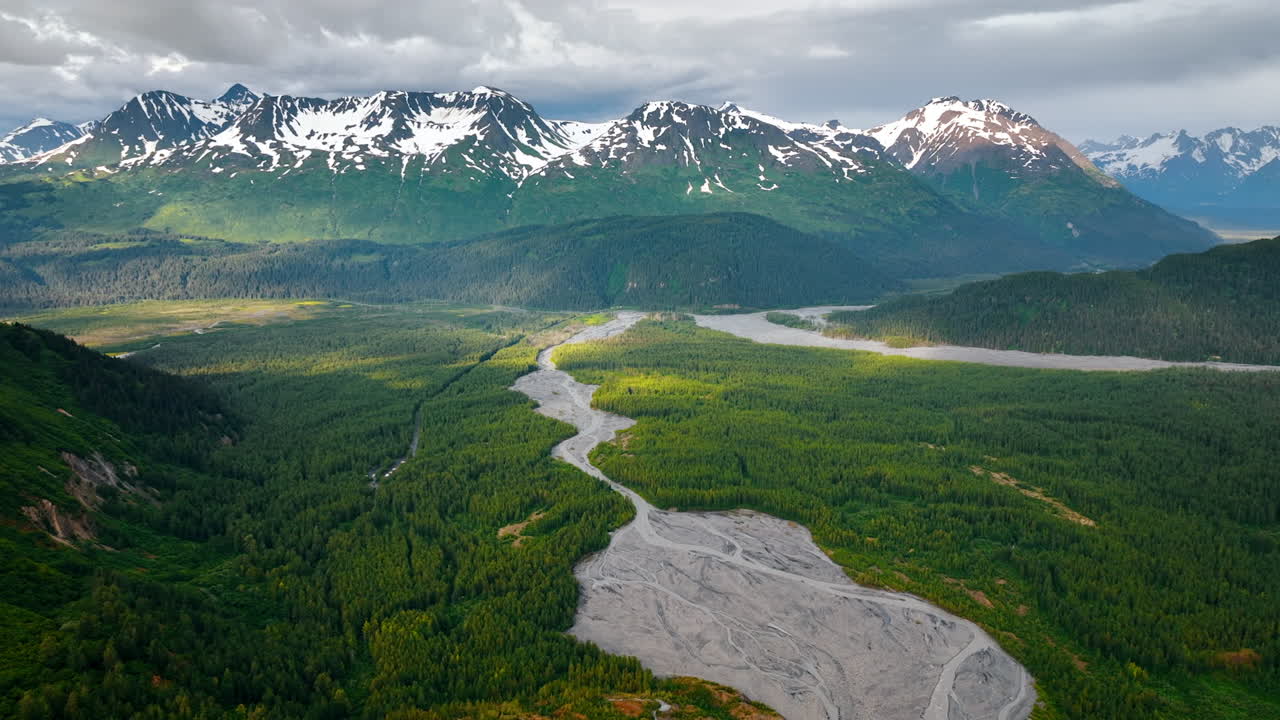 Vihreät metsät peittävät laaksoa, jonka matala joki halkoo. Drone-kuvamateriaali lähestyy upeita lumihuippuisia vuoria pilvisen taivaan alla. Alaska, USA