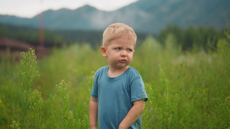 Upset-little-boy-stands-among-high-green-grass-in-lush-field