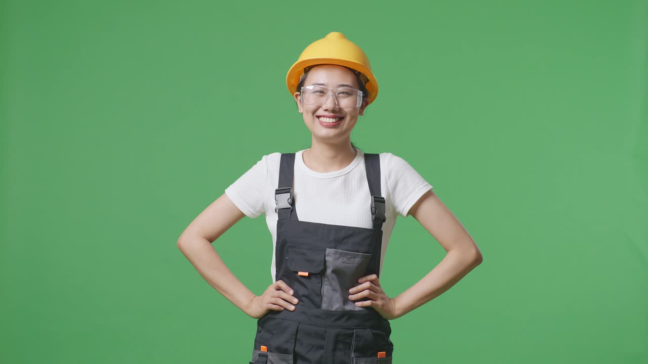 trabajadora asiática con gafas de protección y casco de seguridad de pie con los brazos akimbo mirando a la cámara y sonriendo en el estudio de fondo de la pantalla verde