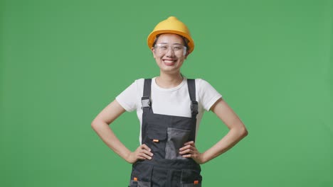 asian woman worker wearing goggles and safety helmet standing with arms akimbo looking at camera and smiling in the green screen background studio