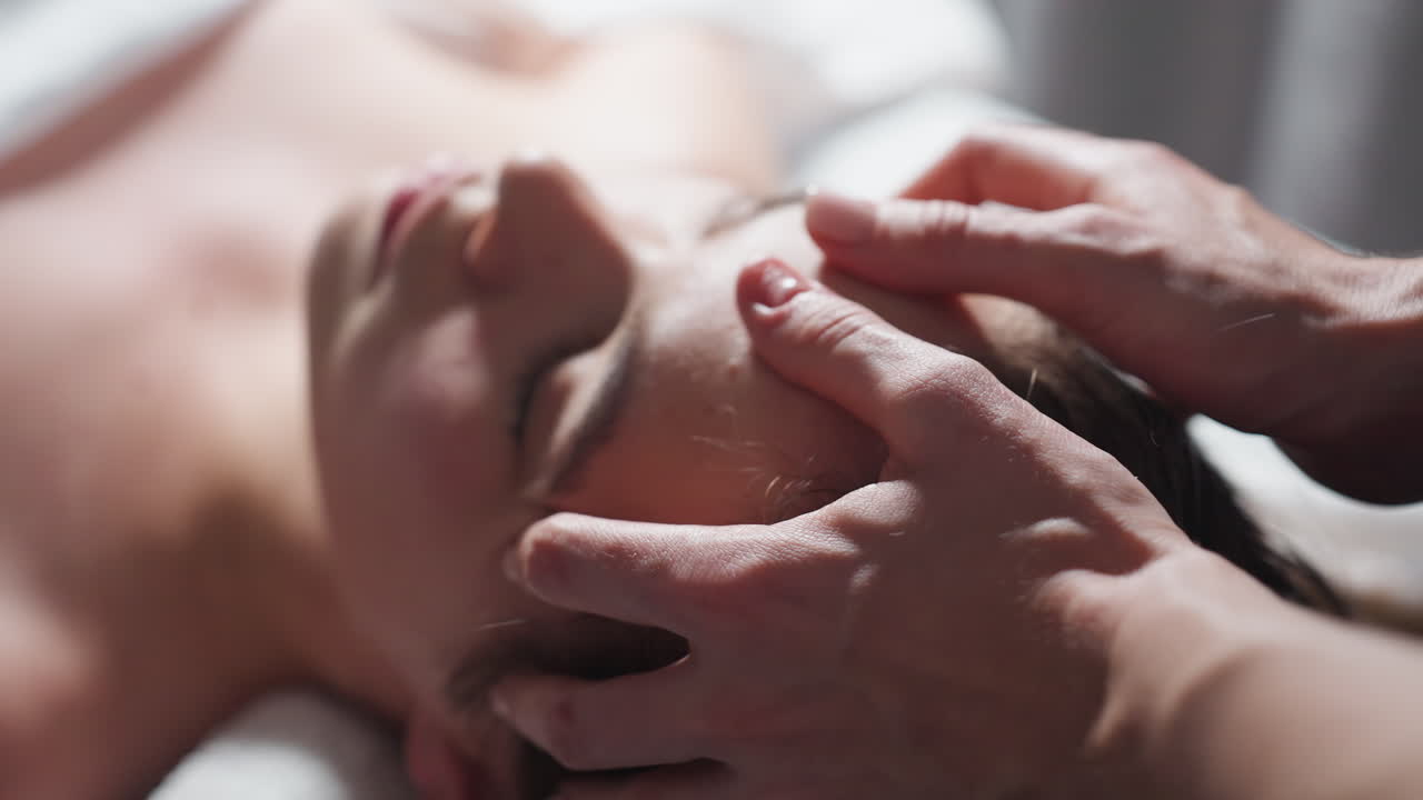 Close up view of woman lying with eyes closed while therapist gently massages her forehead using both hands, capturing serene moment of relaxation and care in peaceful spa treatment environment
