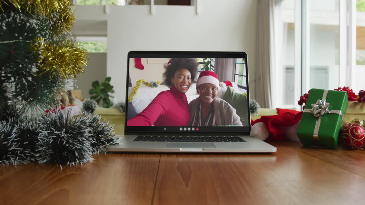 sonrientes madre e hija afroamericanas con sombreros de santa claus en una videollamada de navidad en una computadora portátil