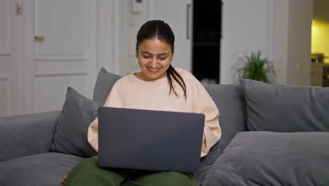 Happy-brunette-girl-in-a-beige-sweater-sits-on-a-gray-sofa-and-works-online-at-her-laptop-in-a-modern-apartment-during-the-day