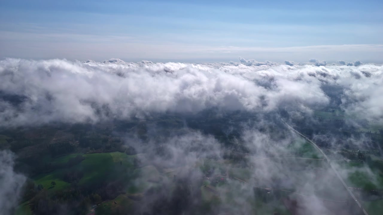 Penerbangan maju udara yang dramatis melalui lapisan awan rendah yang bergejolak dengan celah gelap yang menampakkan hutan hijau dan ladang di bawahnya di bawah langit biru di atas