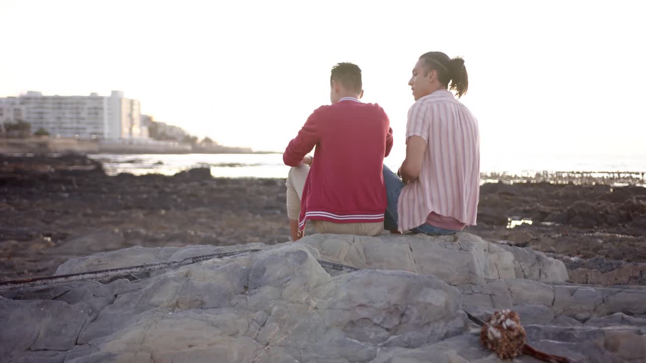 feliz pareja masculina gay diversa sentada en rocas y abrazándose en la playa, cámara lenta