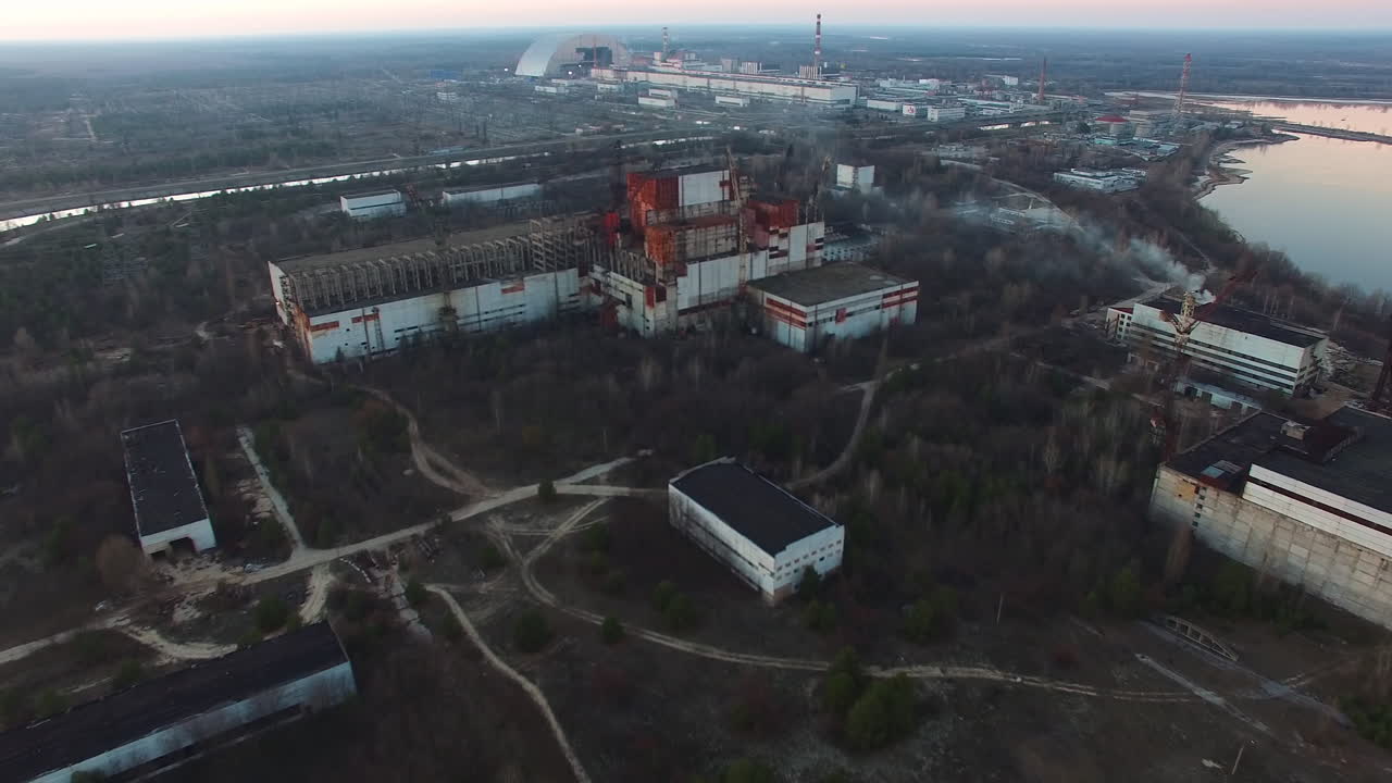 Rozległy widok lotniczy na Czarnobylską Elektrownię Jądrową. Niedokończone, opuszczone bloki reaktorów 5 i 6 wykazują oznaki rozkładu. Nowa Bezpieczna Osłona jest wyraźnie widoczna.