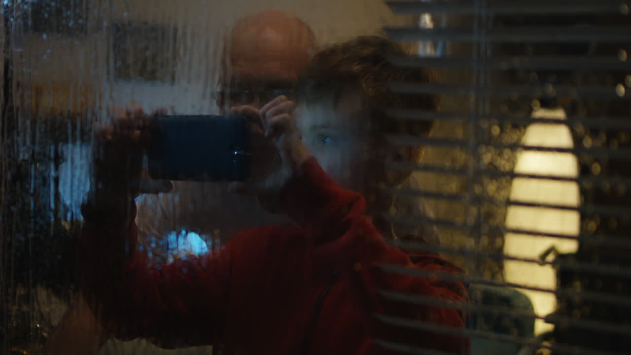 A boy and his father take pictures at night through a rainy window