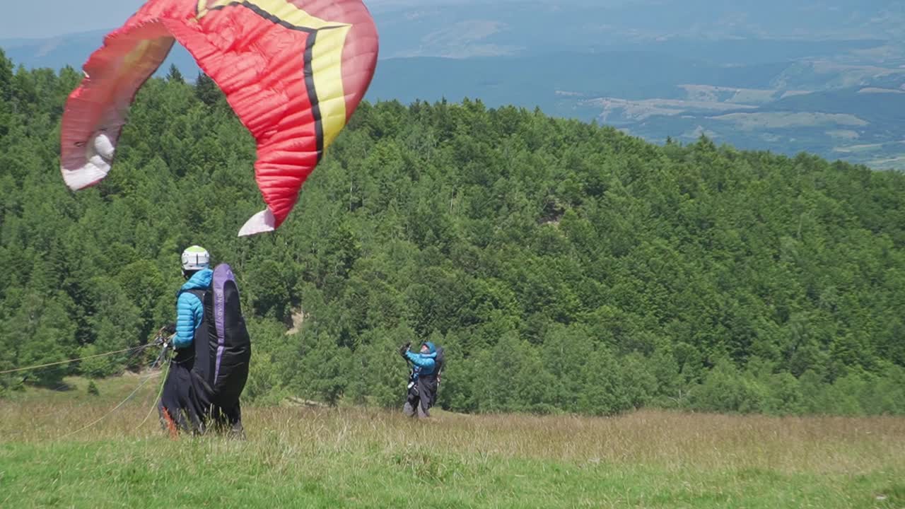 A paraglider ensures his canopy is in perfect position before running forward and lifting off, with a fellow adventurer observing.