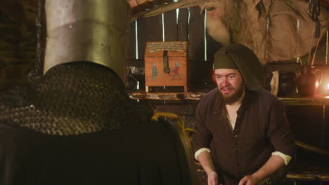 a knight in full armor stands in a medieval setting, a bearded man in a hat sits behind a table.