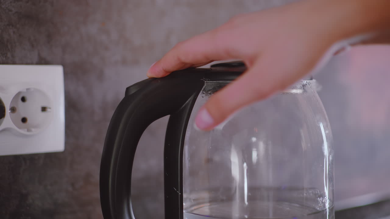 Close up hand pressing button on electric kettle filled with water, kitchen appliance plugged to socket against wall background, preparing to boil water for tea