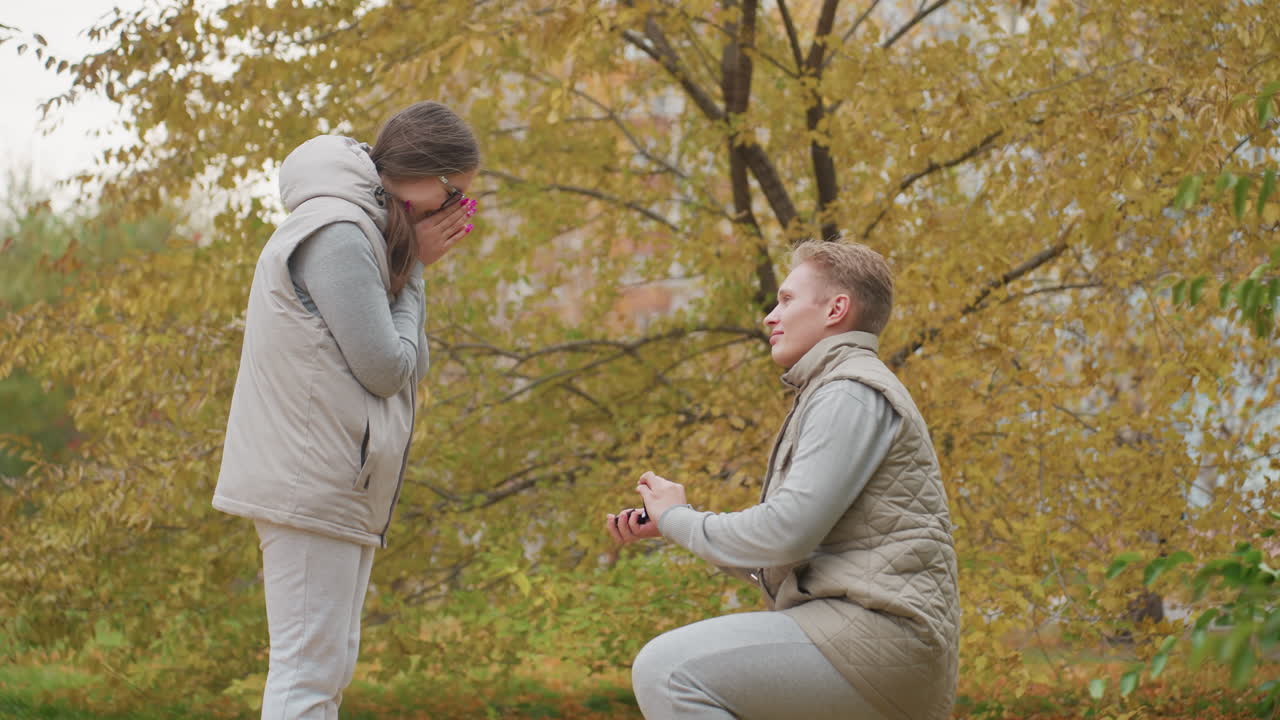 Adults dressed in matching outfits stand outdoors during autumn as man kneels to propose woman reacts by covering mouth in surprise golden leaves sway in wind