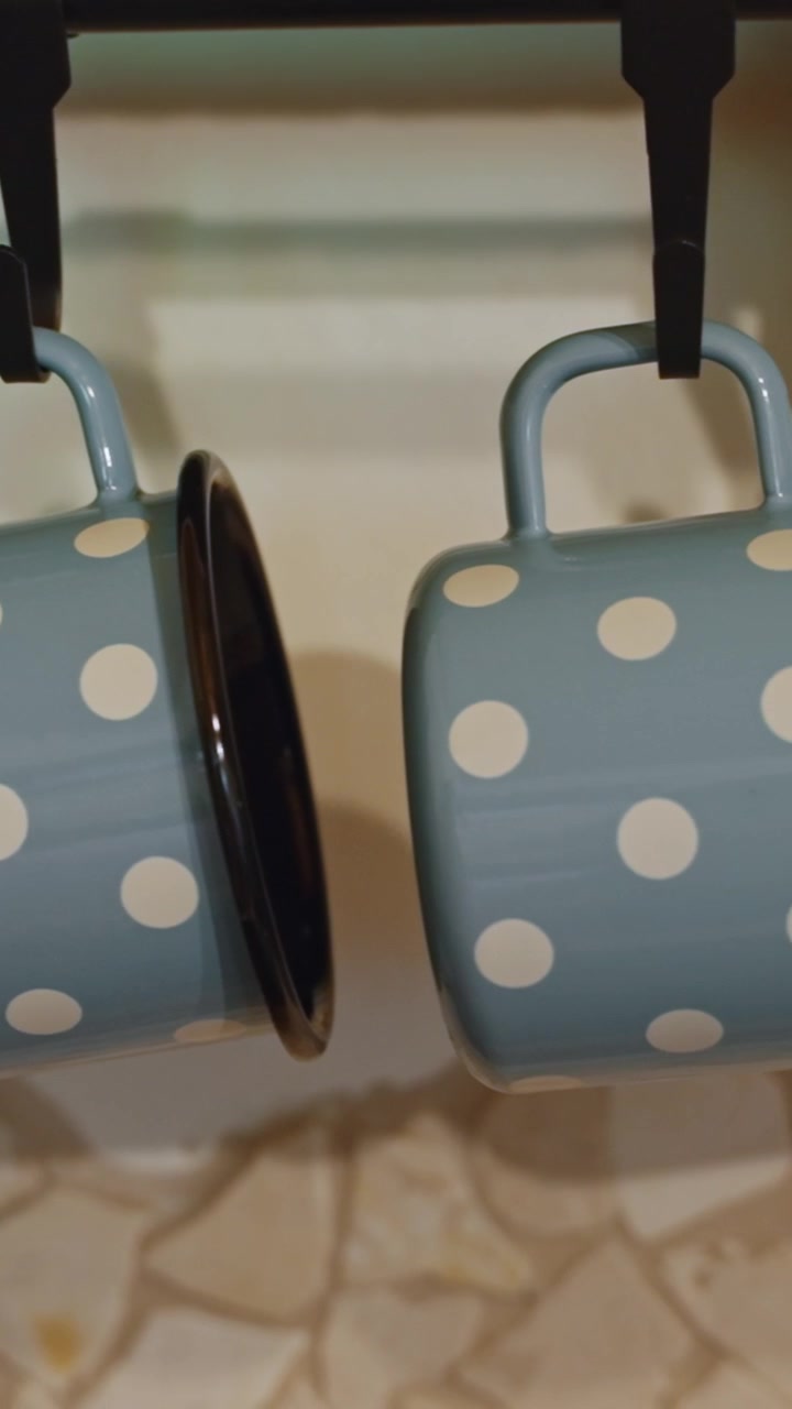 Charming close-up of a blue enamel mug with white polka dots hanging on a kitchen rack. Warm lighting and vertical video, ideal for mobile use.