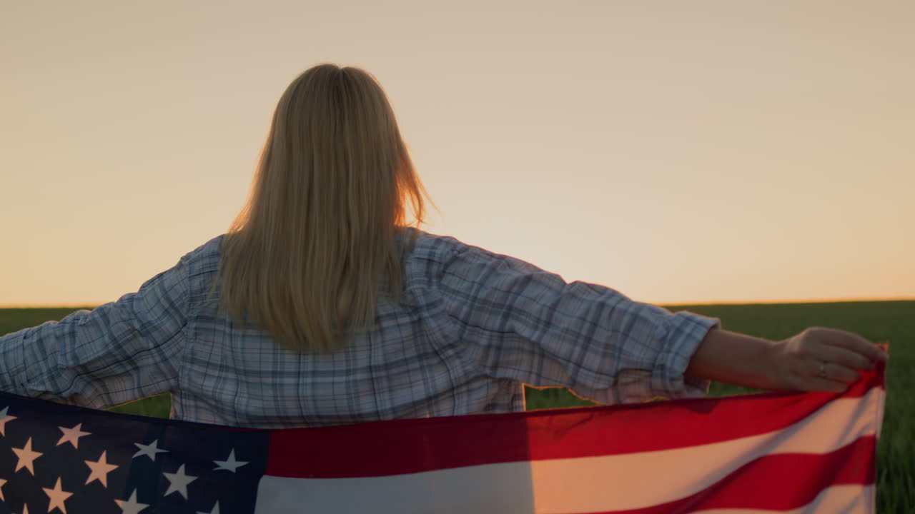 una mujer con una bandera estadounidense en los hombros mira el atardecer sobre un campo de trigo.