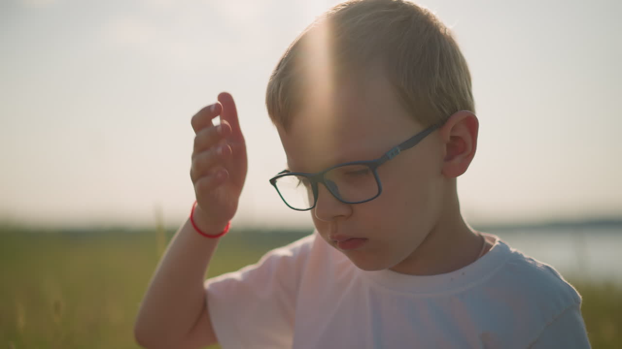 A young boy wearing a white top and glasses, with a red band on his wrist, is sitting in a grassy field. He gently touches his head and appears sleepy, bathed in soft sunlight