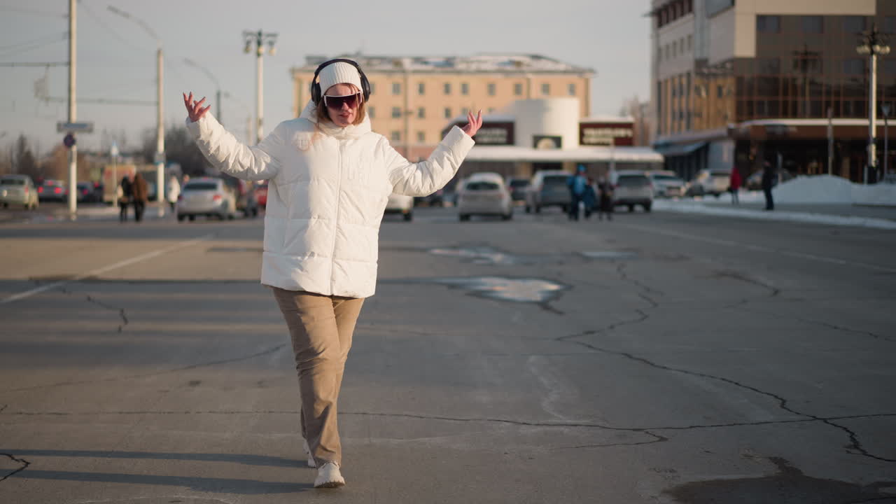 Joyful young lady dances on crack pavement and sings with headphones on, wearing white winter jacket and beanie, enjoying music while moving across sun snowy pavement in afternoon