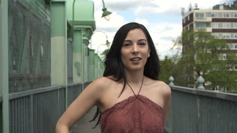 slow motion of an attractive and playful latina woman with black wavy hair and a jean jacket walking on a bridge in london, looking at the camera, happy with a beautiful smile