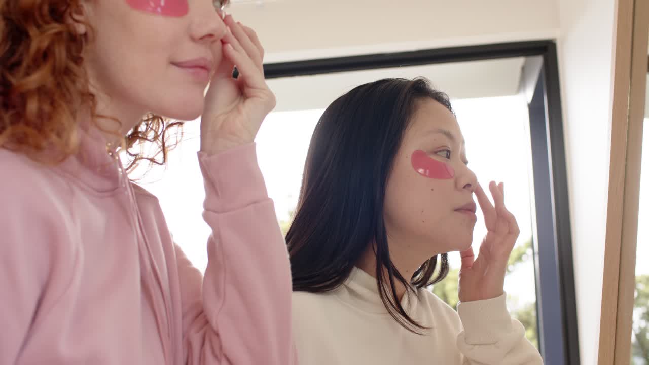Diverse female friends applying pink hydrogel eye patches while checking mirror after seeing friend