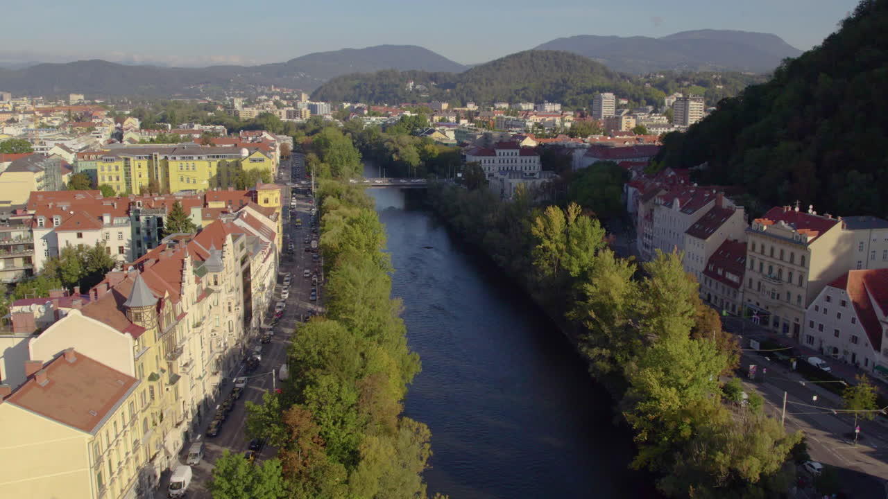 Luftfoto langs den naturskønne flod Mur med livlige røde tagbygninger langs bredderne af Schlossberg-bakken, Østrig