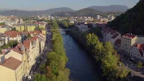 Aerial-view-following-scenic-river-Mur-vibrant-red-rooftop-buildings-along-the-banks-of-Schlossberg-hill,-Austria