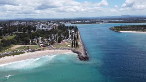 Drone-ascending-over-the-ocean-showing-a-sandy-beach-and-a-river-with-a-coastal-town-in-the-background