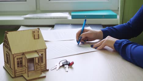 una mujer firma un contrato de vivienda. compra de bienes raíces. hay una casa de madera en la mesa, las llaves de la propiedad