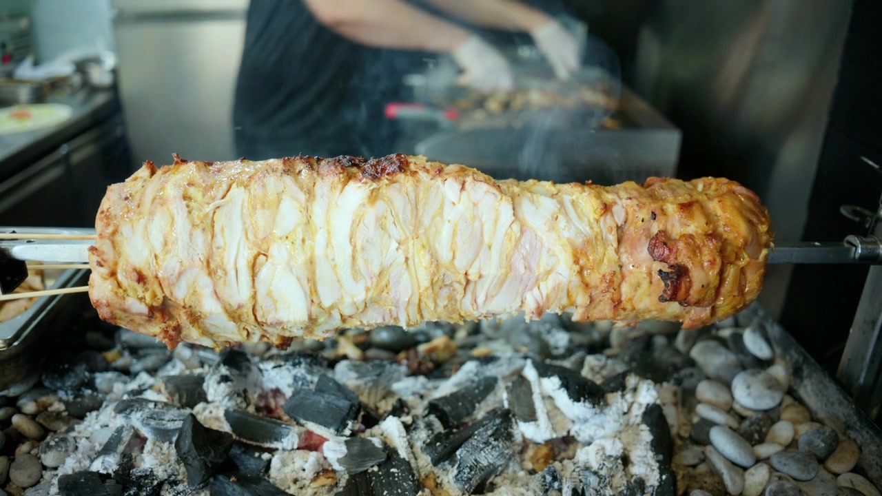 Mano con guanto che affetta succulenta carne di shawarma da uno spiedo verticale per girarrosto, che arrostisce su braci ardenti e pietre.