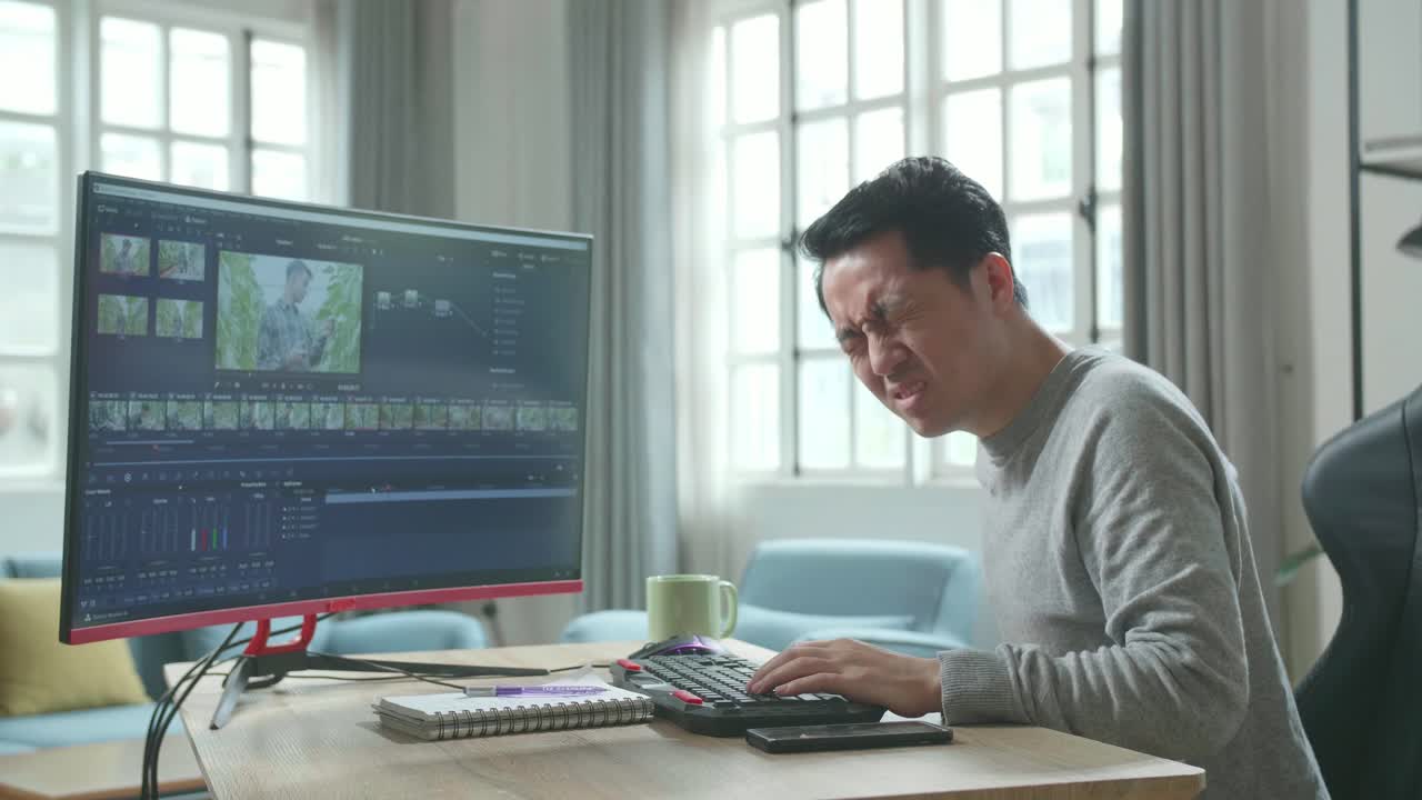 Asian Video Editor Man In Long Sleeved T-Shirt Having Backache While Using Desktop Computer For Editing Video At Home.