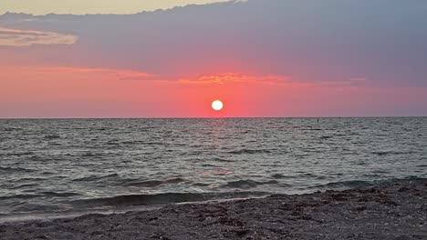 the sun sets across calm waves and a soft sandy beach, coloring the sky in pink and orange hues as reflections shimmer on the tranquil sea surface.