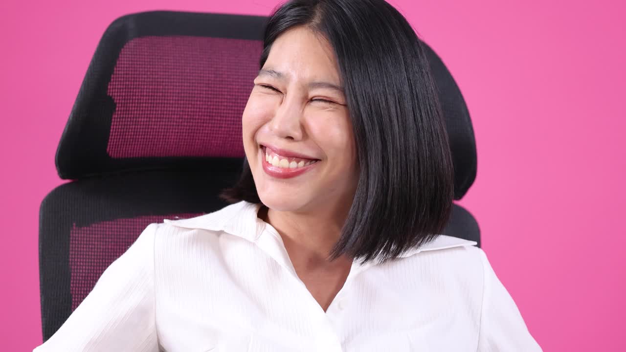 Young Asian woman laughing and gesturing expressively in modern office chair against pink background