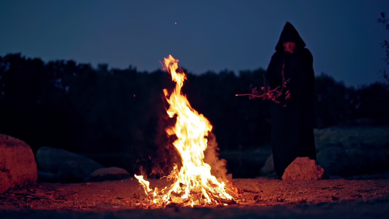 Witch in black robe doing ritual near the bonfire. Vivid fire and sorcerer. Mysterious dark figure conjuring outdoors. Halloween.