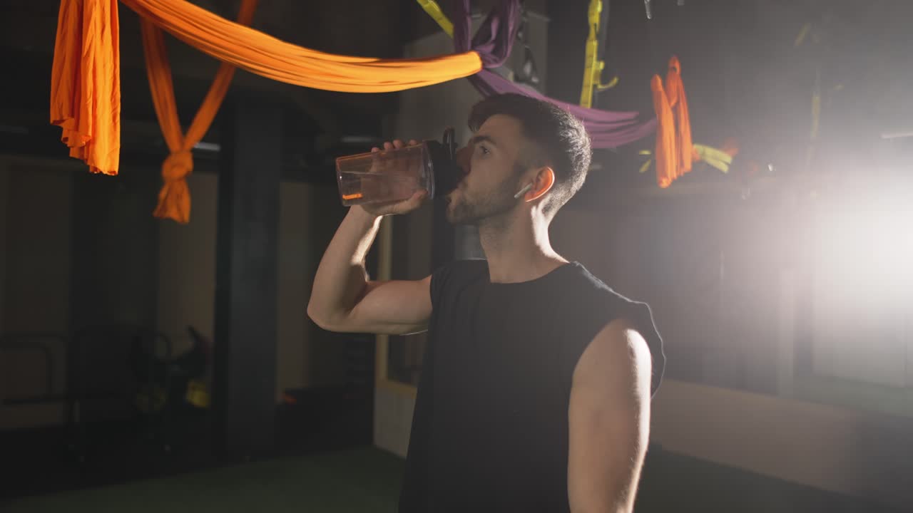Uomo robusto che beve acqua dalla bottiglia in una palestra buia.