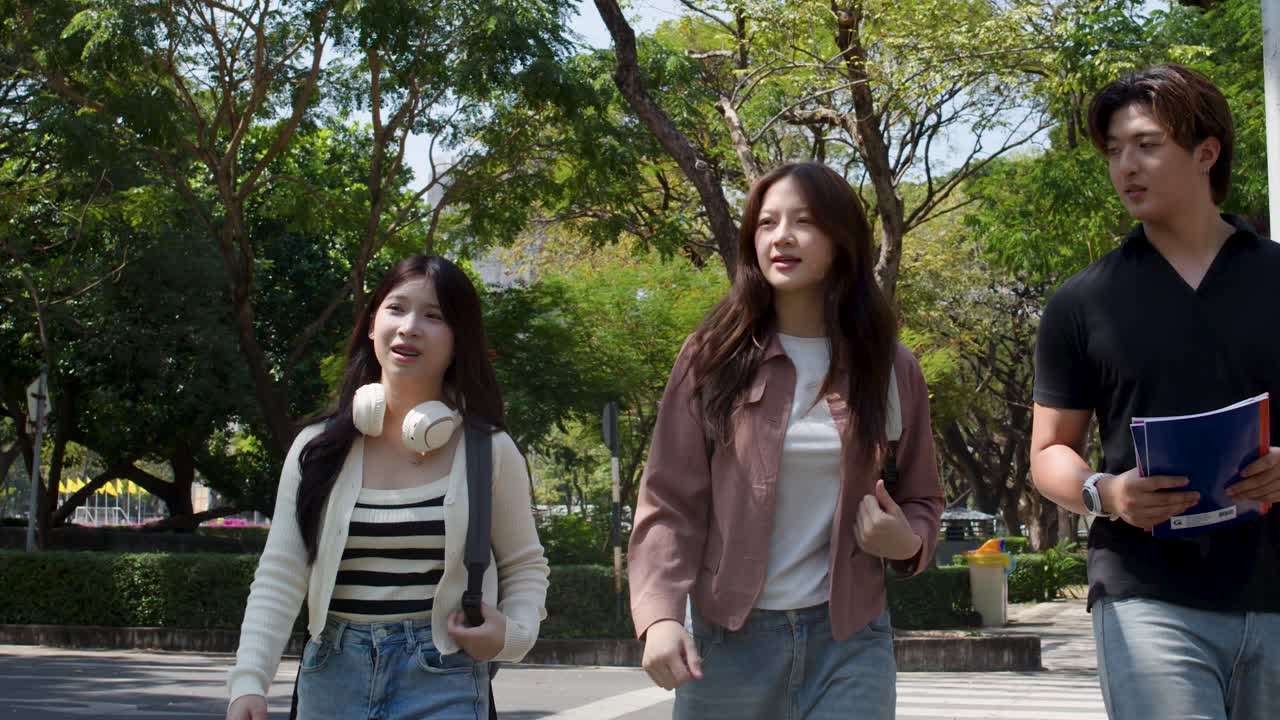 นักเรียนชาวเอเชียสามคนเดินข้ามวิทยาเขตที่แดดจ้าขณะพูดคุยและถือสมุดบันทึกกลางแจ้ง