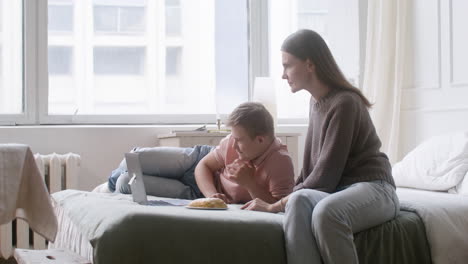 boy with down syndrome and his mother watching something on laptop lying on the bed in the bedroom at home