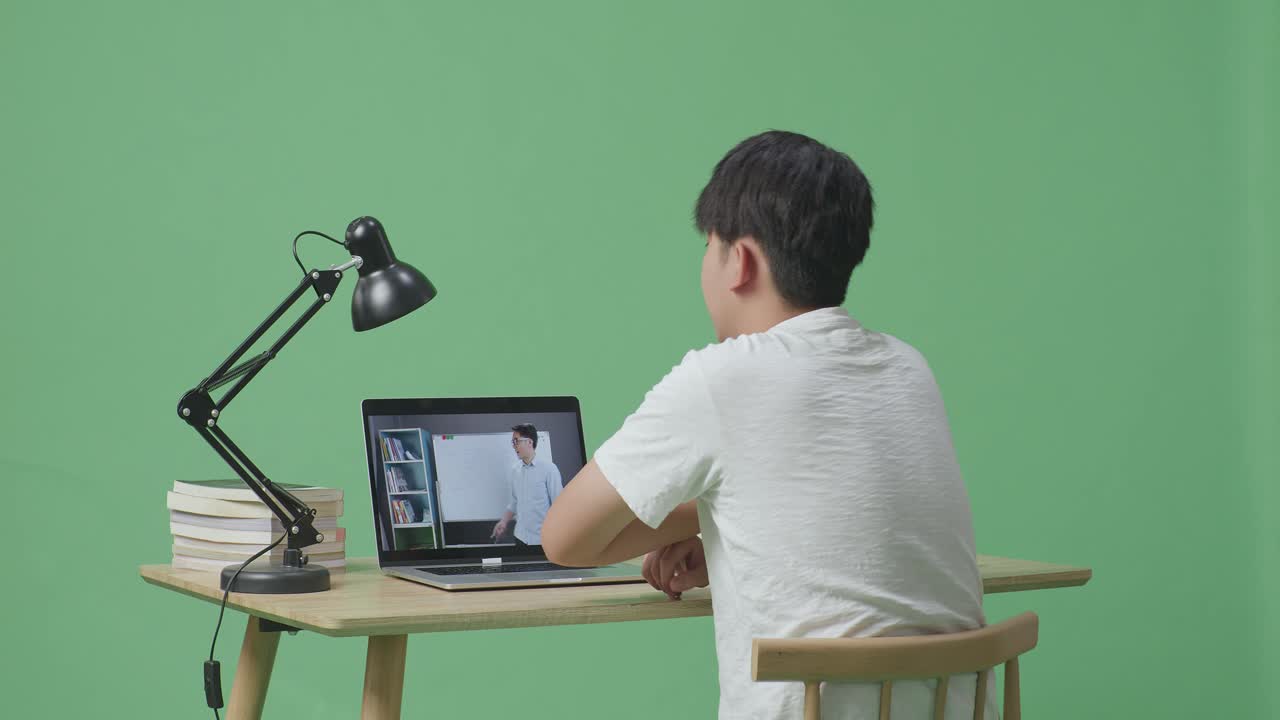 젊은 아시아 남성이 녹색 스크린 배경에서 공부하는 동안 질문을 하기 위해 손을 들고 집에서 노트북으로 가르치는 남성 교사를 보고 있습니다.