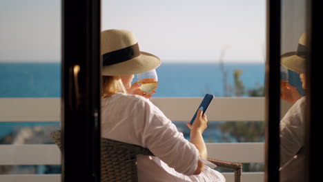 frau mit weinglas benutzt eine zelle, wenn sie sich auf dem balkon mit blick auf das meer entspannt