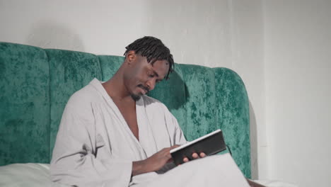 man in white bathrobe sitting on bed with green velvet headboard, holding black notebook, smiling softly while reading, relaxed atmosphere, cozy indoor setting