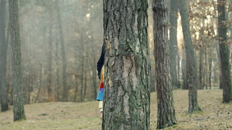 Portrait-shot-of-Caucasian-handsome-dad-and-his-cute-little-boy-and-girl-looking-at-camera-behind-a-tree-trunk-in-the-forest