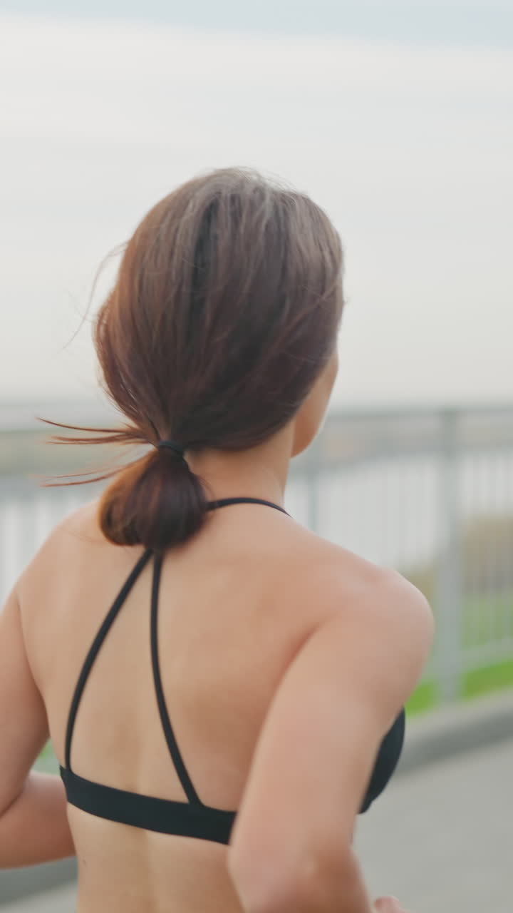 Fair girl in black neck top jogging beside iron fence with blurred view of a woman walking past her in park, capturing outdoor fitness, health, and motion in bright surroundings