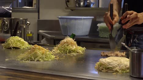 Preparing-traditional-japanese-okonomiyaki-food-with-coleslaw-and-meat,-on-a-steel-area