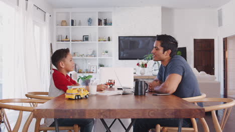Hispanic-father-and-son-talking-while-they-work-sitting-at-the-dining-table,-side-view