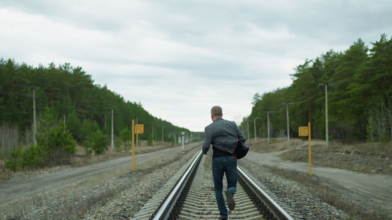 A close view of an aged man running on a railway track, looking back and then forward, he is wearing a grey suit, jeans, and canvas shoes, with trees, electric poles, and two yellow poles