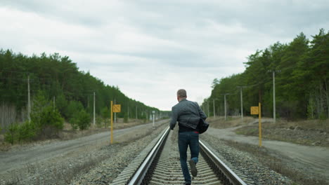 a close view of an aged man running on a railway track, looking back and then forward, he is wearing a grey suit, jeans, and canvas shoes, with trees, electric poles, and two yellow poles