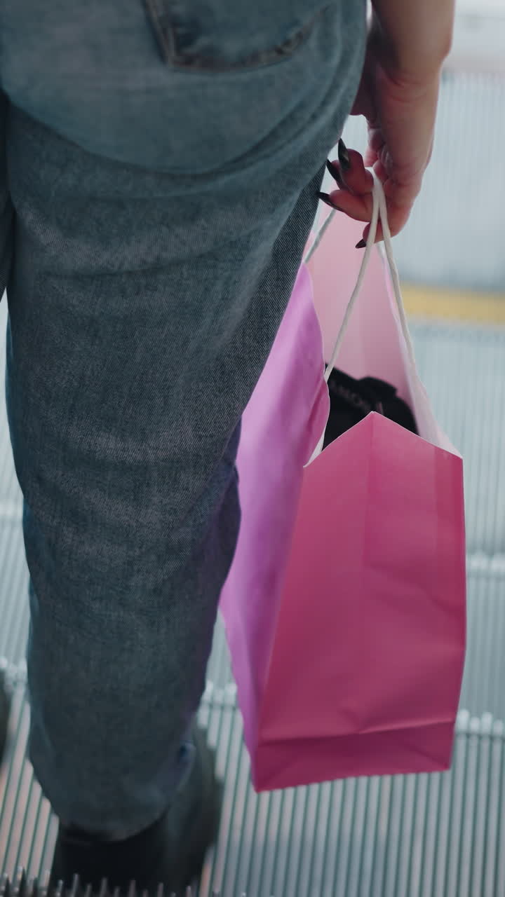 vista trasera de una mujer en vaqueros y botas negras llevando bolsas de compras, bajando de la escalera mecánica y caminando hacia la puerta con un ligero borrón en el fondo