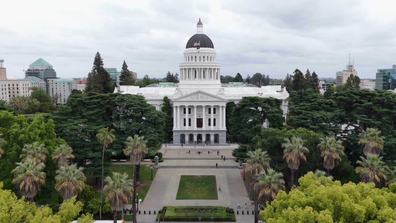 Vista aérea de drone do edifício do Capitólio Estadual da Califórnia em Sacramento, Estados Unidos, com a icónica cúpula branca, bandeira americana e palmeiras circundantes no distrito governamental histórico sob céu nublado
