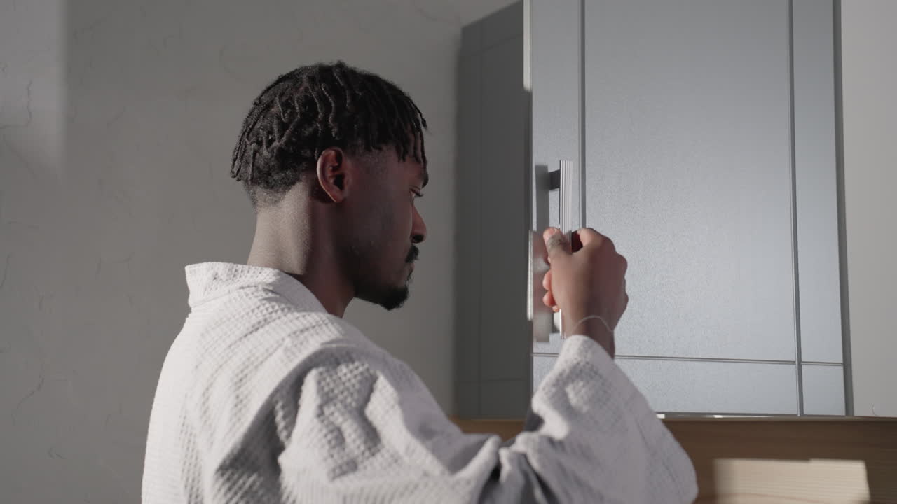 Man in bathrobe opening kitchen cabinet door in modern minimalist kitchen. Soft light highlights his profile and clean kitchen design, with shadows cast on the wall
