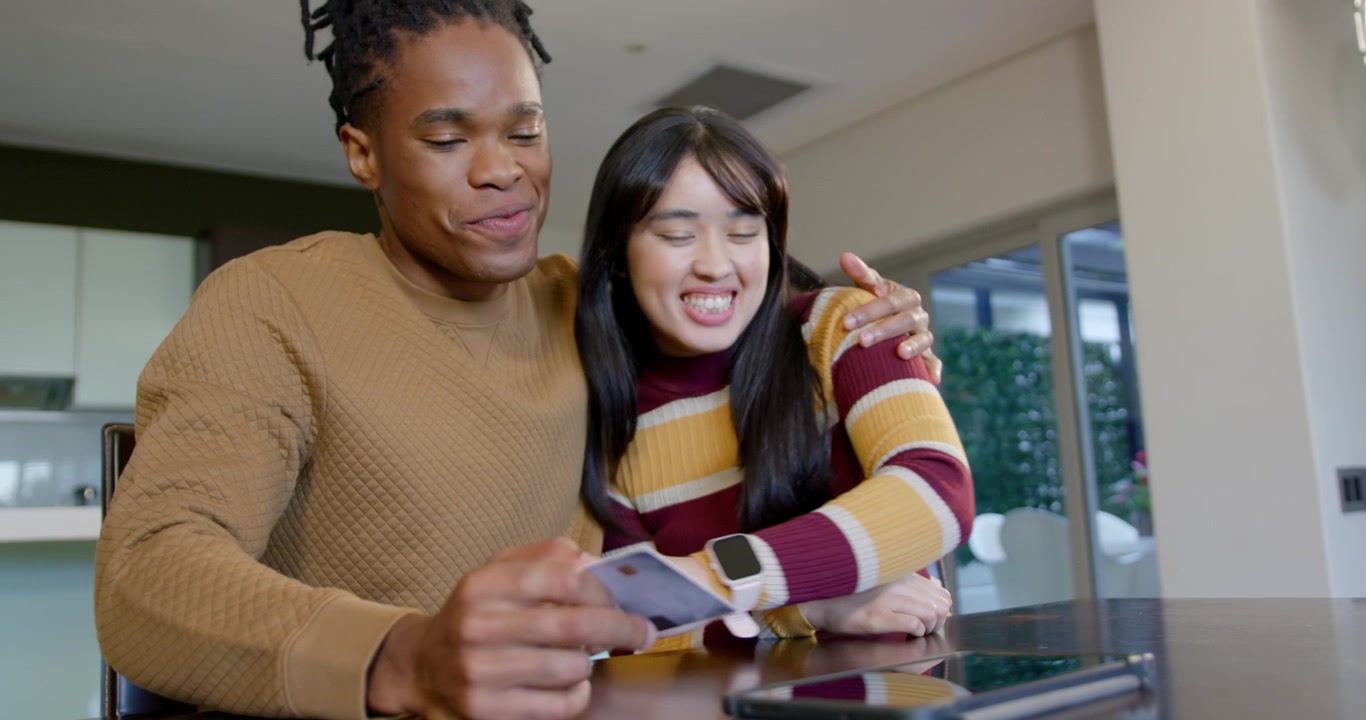 Diverse couple at dining table, man showing payment card to tablet folio case to pay, hugging
